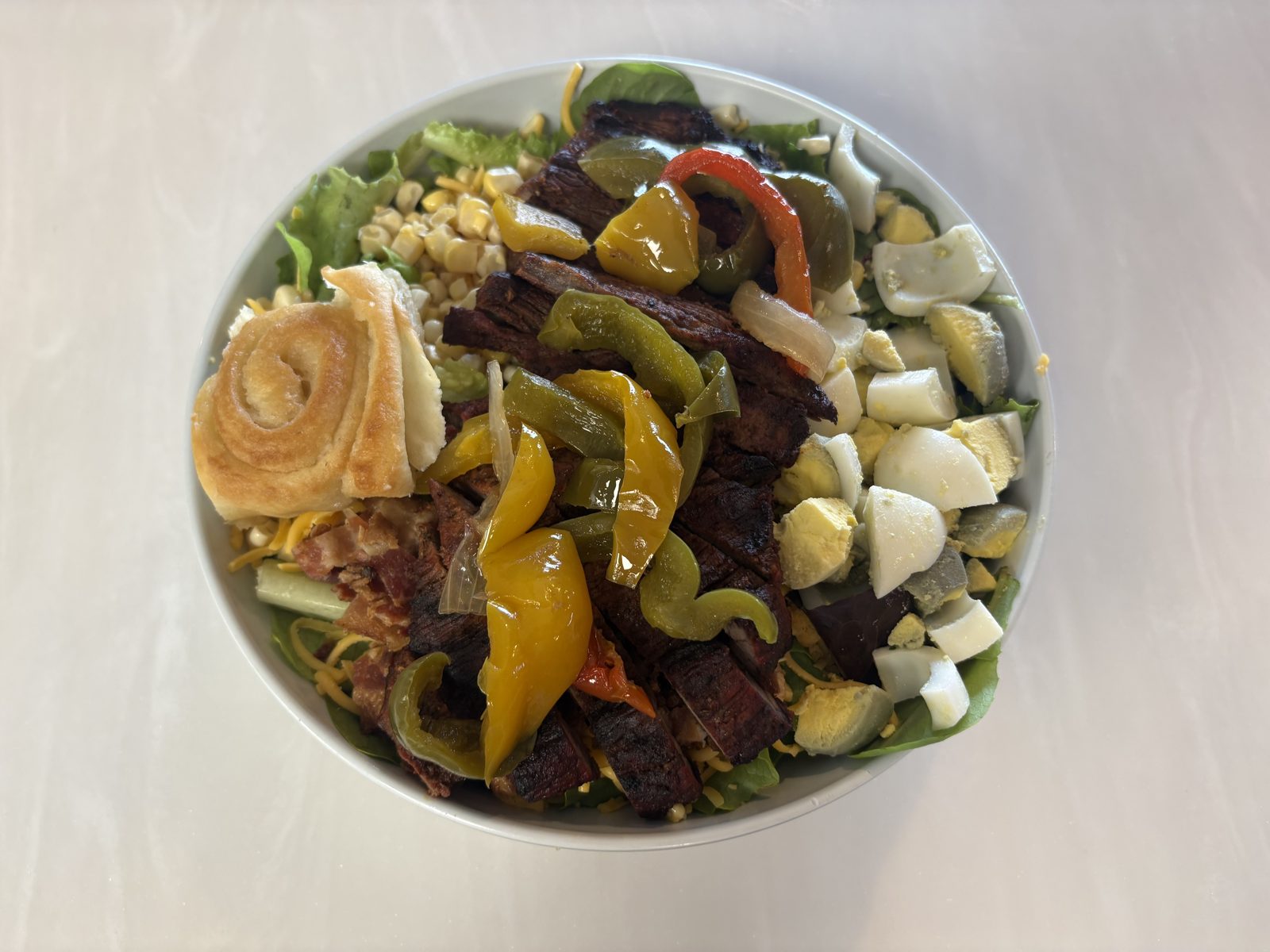 A salad topped with sliced steak, peppers, boiled egg, corn and cheese